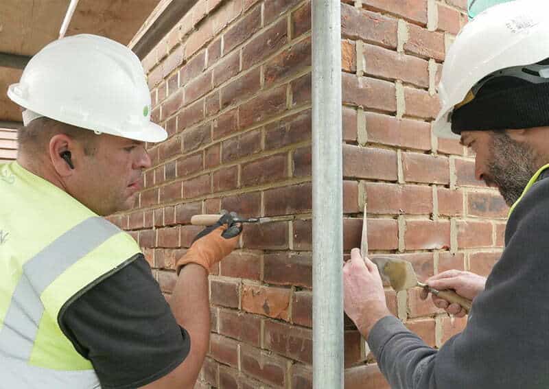 Two construction workers in hard hats and safety gear repairing a brick wall, demonstrating professional damp-proofing techniques for moisture protection.