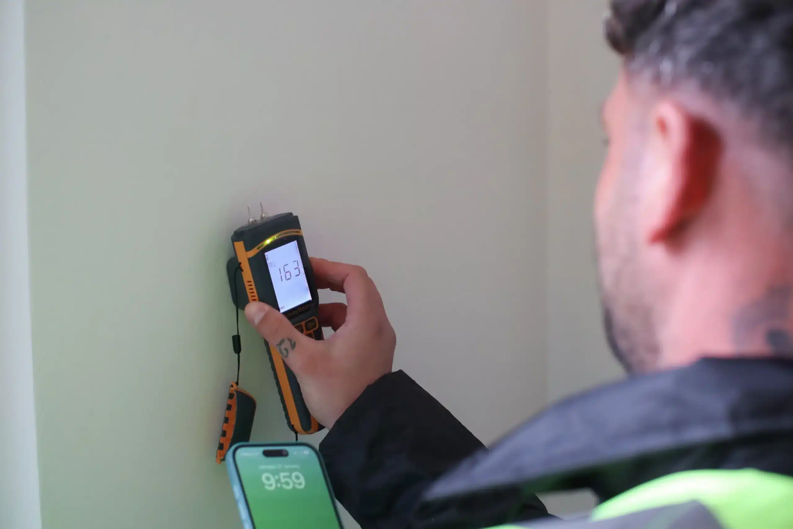 Person using a damp meter to measure moisture levels on a wall, with a smartphone displaying the time in the foreground, relevant to professional brick cleaning and sealing services.